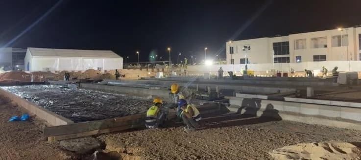 Night shift crew of about five in hi-vis and helmets crouched fixing timber shuttering along kerbed bays; tent and building lit behind