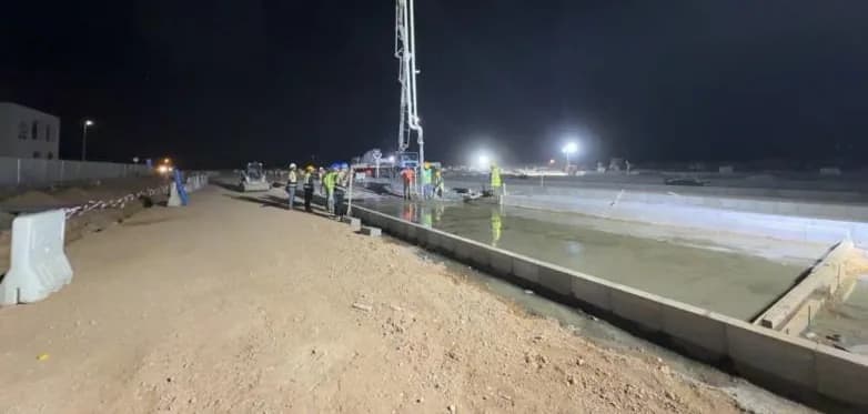 Night pour: concrete pump boom over freshly placed CLSM in a kerbed bay, crew of about six in hi-vis at mid distance