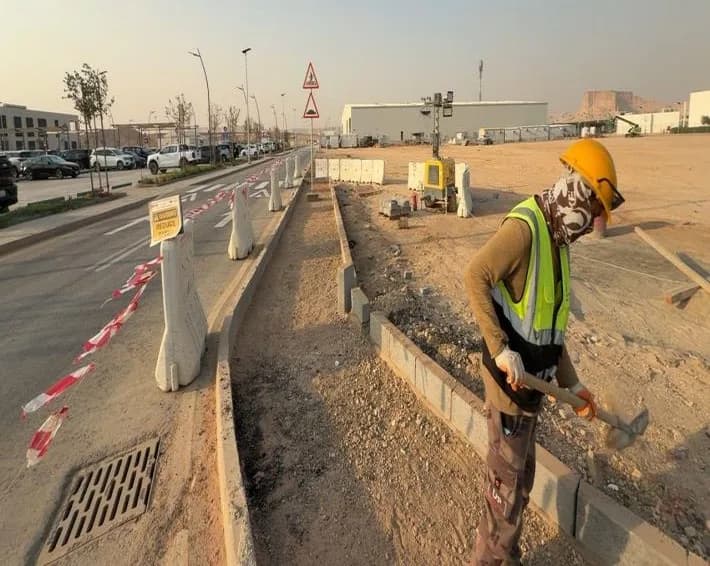Worker with scarf-covered face swinging a pick in a kerb-edged strip beside a barricaded road, red-white tape on barriers, drainage grate, speed hump warning signs and light tower behind