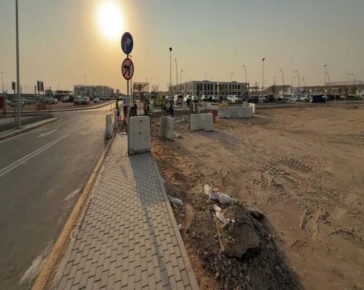 Interlock pedestrian walkway running beside a road toward the low sun, turn-right and no-left-turn signs, barriers, unpaved strip with debris, distant workers and vehicles