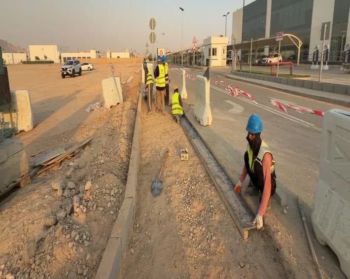 Crouching worker with scarf-covered face setting a steel edge form along a kerb median, other workers behind, barriers with tape, building with FDC sign and vehicles at distance