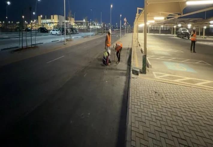 Night view of a finished asphalt road with lane markings, interlock walkway in foreground, lit canopy parking with accessible bays at right, three workers on the road and one under the canopy, lit buildings behind