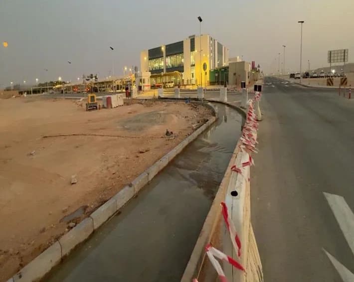 Dusk view along a road with a kerb-edged channel holding standing water, barriers with red-white tape, unpaved area with equipment at left, lit building and distant vehicles