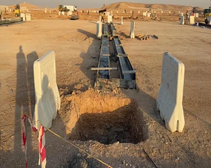 Square excavation in the foreground between two water-filled barriers, steel channel formwork running towards distant workers and plant at sunrise; photographer's shadow at left