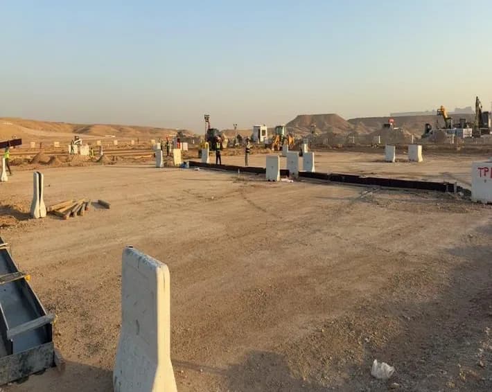 Graded parking area at sunrise with precast blocks and water-filled barriers, a line of dark edge formwork, workers and backhoe loaders in the distance