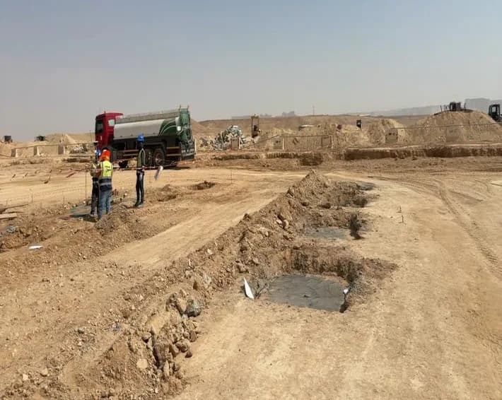 Row of square foundation pits with concrete bases in the foreground, three surveyors in hi-vis setting out at left, water tanker truck behind