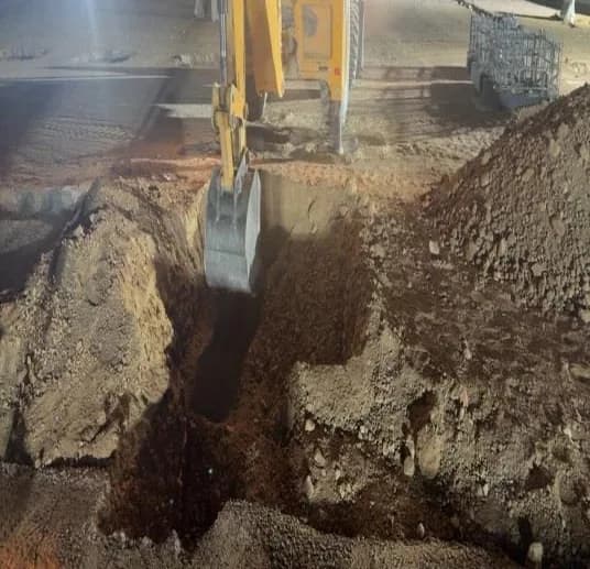 Backhoe loader bucket excavating a square foundation pit at night under floodlights, spoil heaps around, steel mesh cage at right