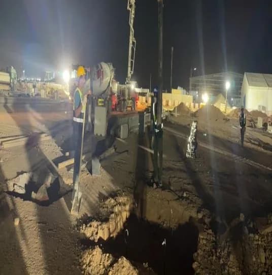 Concrete pump truck with boom raised over a foundation pit at night, worker in hi-vis at left, site buildings lit in the background