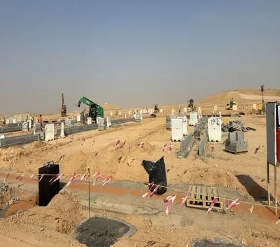 Parking area with precast blocks, low block walls and barrier tape, black plastic-covered pit in the foreground, excavators and loader in the distance