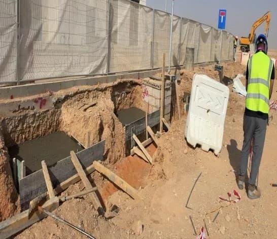 Row of foundation pits with timber formwork and fresh concrete along the fenced site boundary, worker in hi-vis with back to camera, Qiddiya-signed building and No Parking sign behind