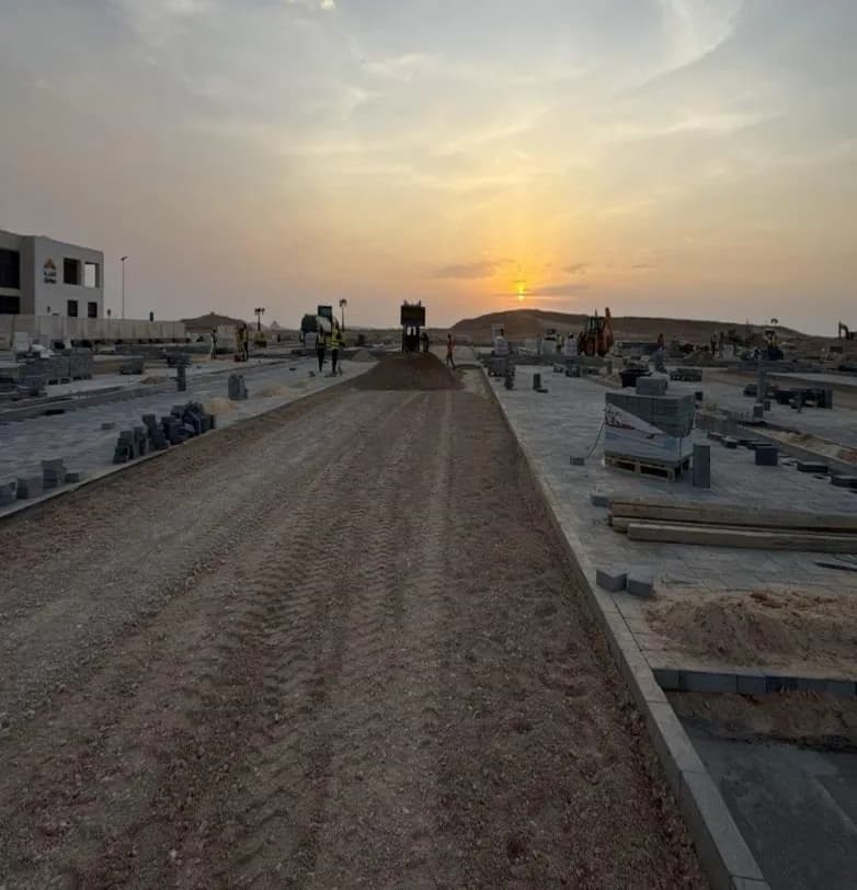Sunset down the access road with aggregate base course spread between interlocked bays, pallets of blocks, roller and workers far away