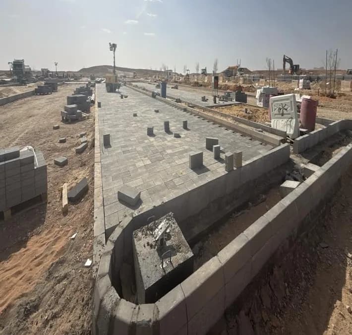 Completed interlock paving on a kerbed island with a lighting pole base and anchor bolts in foreground, pallets of blocks, excavators in distance