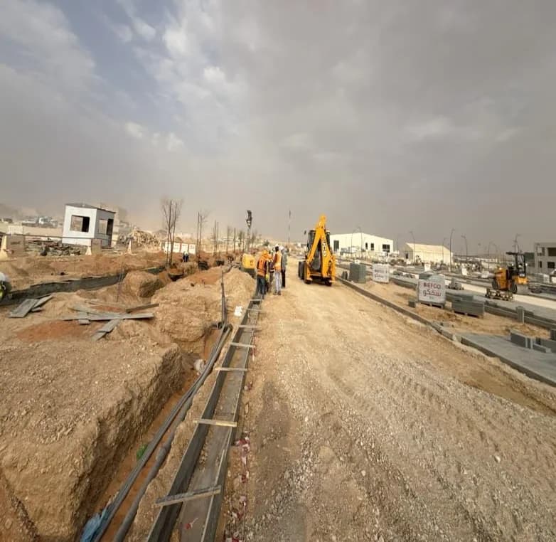 Trench with black pipes and timber edge along the haul road, JCB backhoe, group of four workers in orange vests, SIEFCO bags and paver stacks at right