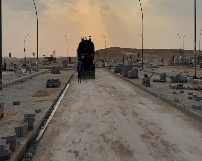 Bitumen distributor truck seen from behind at sunset with a worker hand spraying MC1 prime coat onto the base course along the road corridor; kerbs and paver pallets both sides