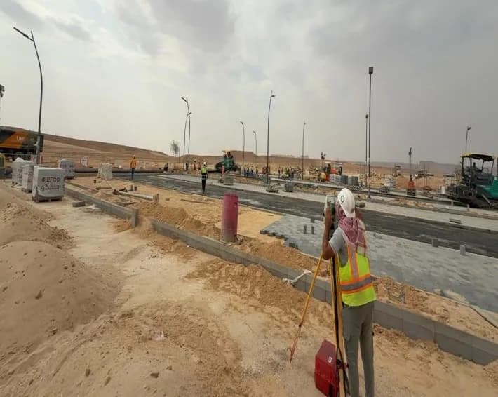 Surveyor in orange vest and white hard hat with red shemagh at a total station, back to camera; sand piles, kerbed bays, asphalt paver and workers distant, IEFCO pallet at left