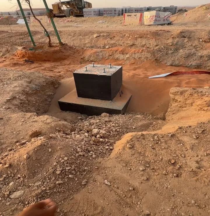 Concrete pedestal foundation with four anchor bolts on a stepped base inside an excavated pit in red sand, staked sapling left, motor grader and SIEFCO block wall behind, photographer's fingers at bottom-left corner