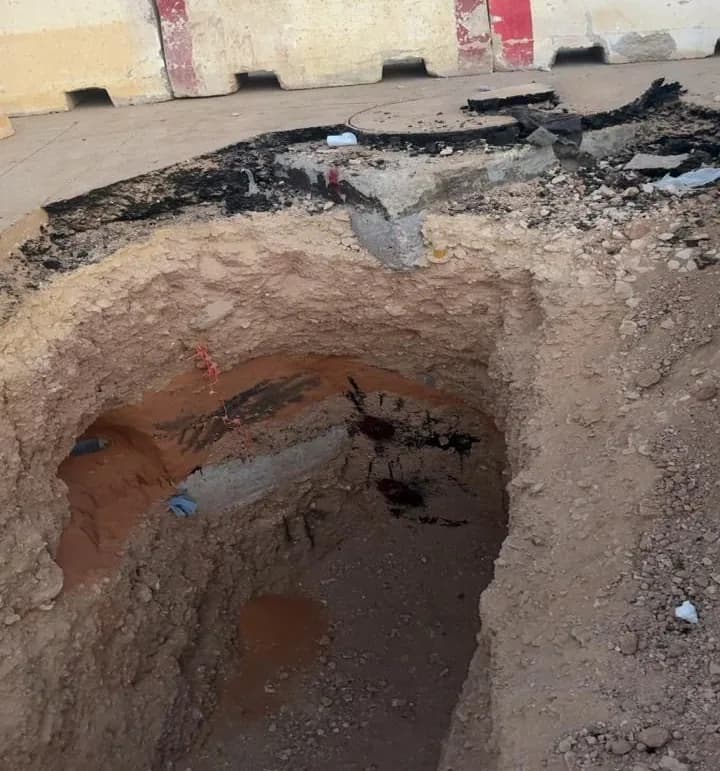Excavated pit through pavement exposing a buried grey drainage pipe, broken asphalt and concrete at the edge, UNIMAC-branded concrete barriers behind