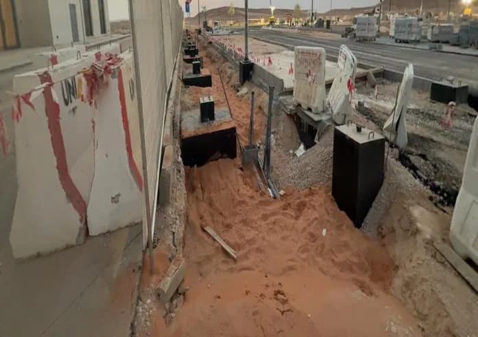 Black-coated concrete pedestal foundations in a trench along a mesh fence, Qiddiya-logo building left, UNIMAC barriers, red sand, road and SIEFCO blocks beyond under sunset sky