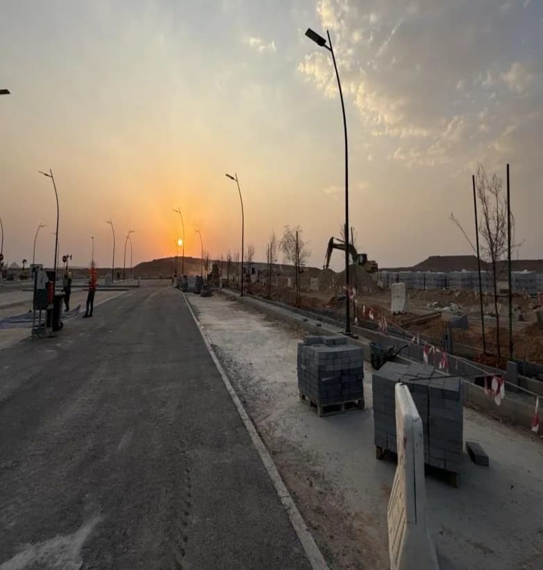 Sunset along access road with interlock pallets on right, planted trees, excavator in distance, two distant workers at left