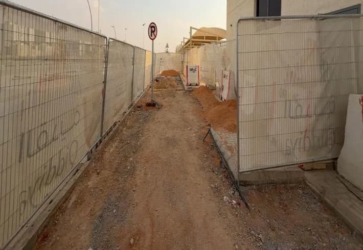 Narrow corridor between mesh fences printed with Qiddiya branding along building wall, excavated soil piles, no-parking sign, shade canopy beyond