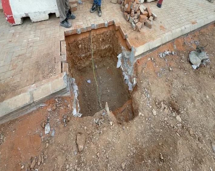 Rectangular pit excavated through interlock walkway with tape measure hanging in, removed blocks stacked, two workers' legs at top, fire extinguisher