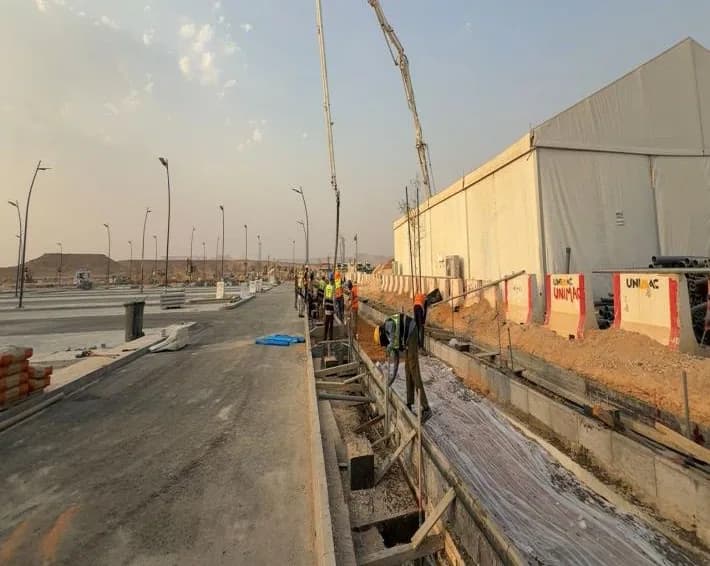 Concrete pump boom extended over the trench with formwork beside the tent; workers at a distance, UNIMAC-branded barriers, paved parking at left