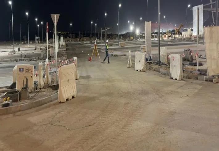 Night view of a surveyor at a total station on a tripod in the roadway between barricades, rows of light poles receding