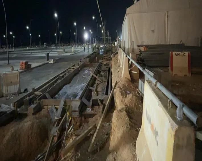 Night view of the concrete pump boom over the kerbed drainage channel with fresh concrete, formwork and stakes; tent and UNIMAC barriers at right, tiny figures in the distance