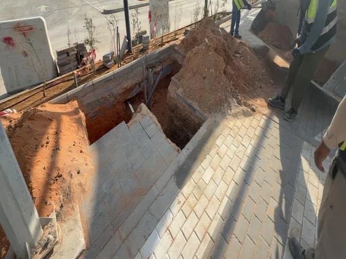 Storm line trench excavated through interlock walkway edge with spoil heaps and pipe visible; three workers; parking canopies behind