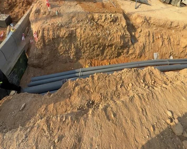 Excavated trench with grey utility conduits running through it beside a kerb line, UNIMAC-marked concrete barrier and stacked pipes at top, site cabins behind
