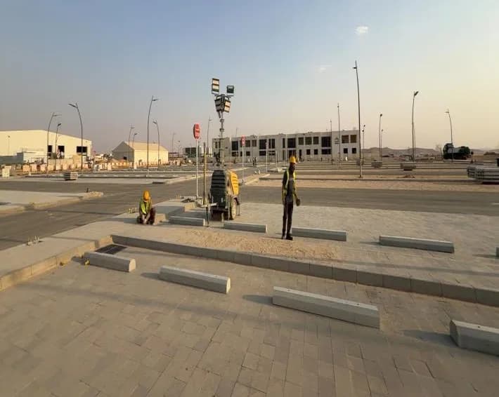 Wide view of interlock-paved parking with concrete wheel stops laid in rows, mobile light tower, two distant workers, drainage grate, buildings and street lighting behind