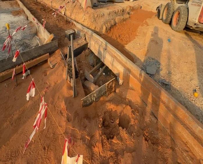 Trench along a kerb-edged line with shutter boards and props, red-white tape, skid-steer loader at right, UNIMAC-marked barriers, tent and cabins behind, photographer's shadow