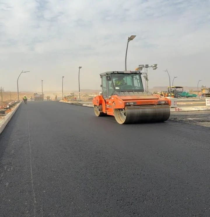 Tandem roller compacting the first asphalt layer. HAMM tandem roller compacting a wide fresh asphalt mat, paver and distant workers at the far end under new streetlights