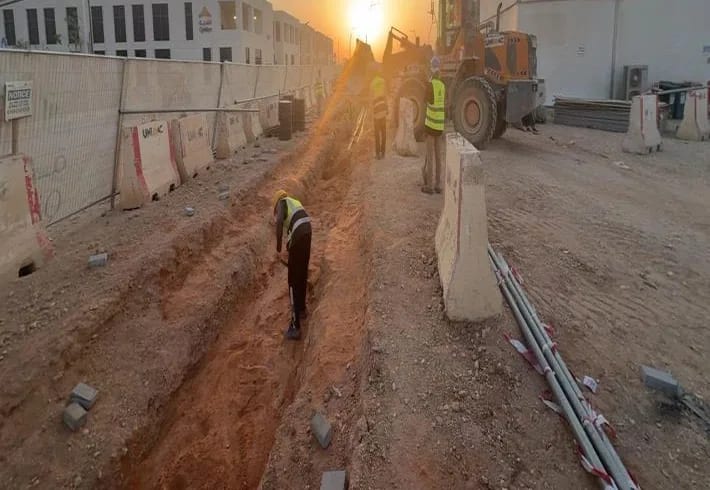 Sunset trench excavation along a fenced corridor, worker digging in trench, UNIMAC wheel loader with three workers beside it, stacked pavers, conduits and barriers, Qiddiya-logo building behind