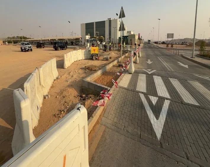 Asphalt road with chevron markings and zebra crossing beside a barricaded kerb median under works, red-white tape, light tower, distant workers and vehicles, glass building behind